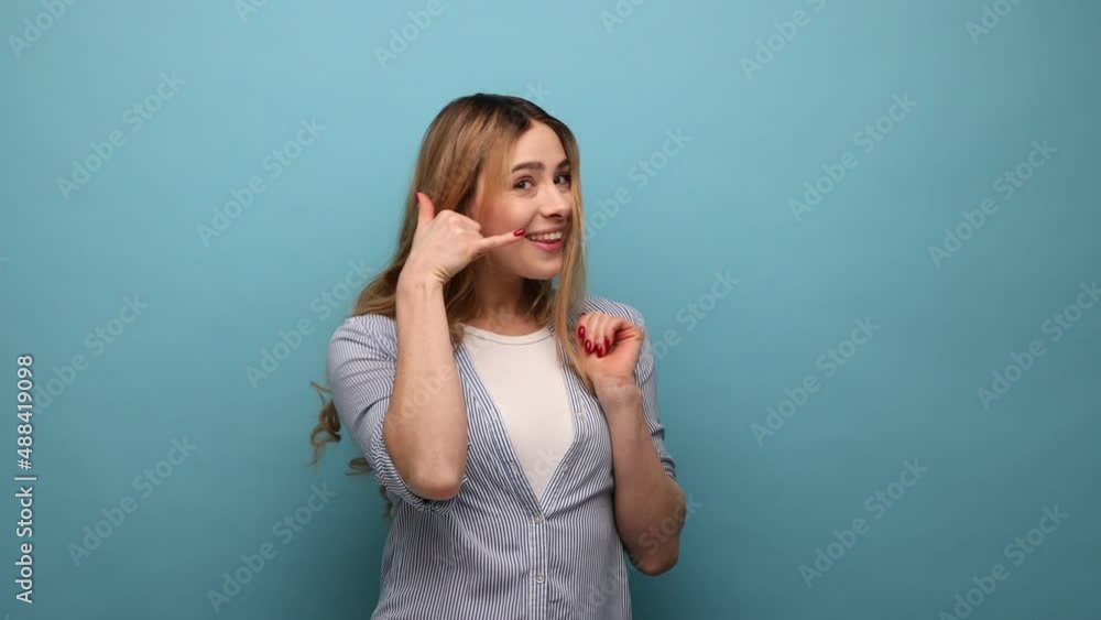 Call me. Woman with wavy hair standing with telephone hand gesture and smiling to camera, flirting offering to contact by phone, wearing striped shirt. Indoor studio shot isolated on blue background. Call me. Woman with wavy hair standing with telephone hand gesture and smiling to camera, flirting offering to contact by phone, wearing striped shirt. Indoor studio shot isolated on blue background.