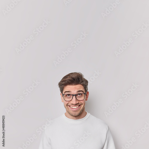 Vertical shot of handsome cheerful European man focused above has broad toothy smile wears transparent glasses and casual jumper notices something isolated over white background blank copy space © wayhome.studio 