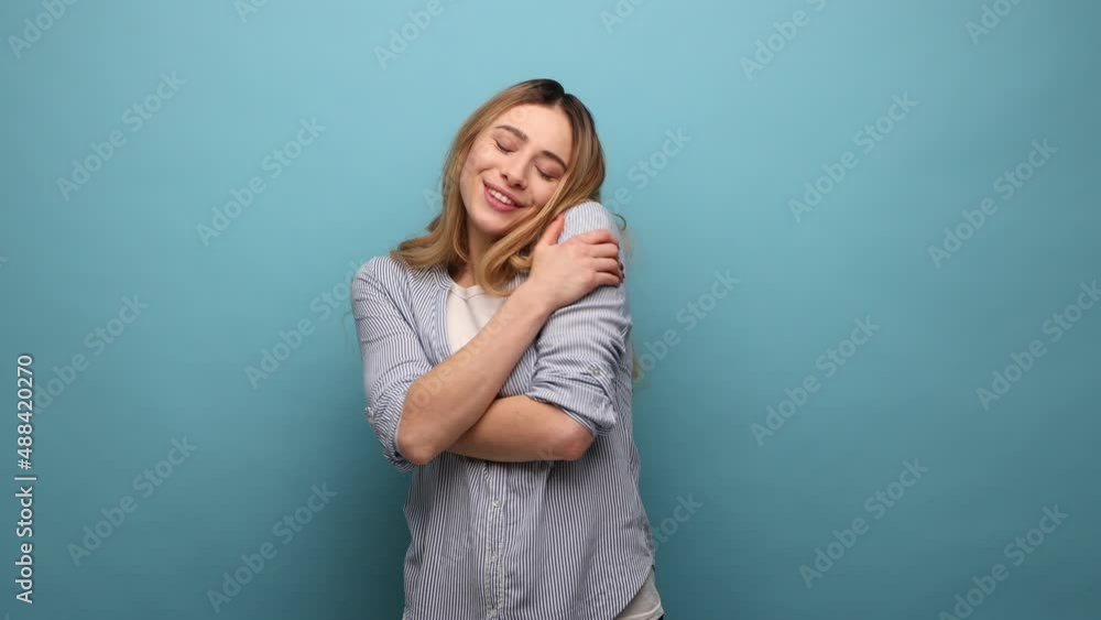 Portrait of beautiful young woman hugging herself tightly and smiling with pleasure, appreciating her appearance, self esteem, wearing striped shirt. Indoor studio shot isolated on blue background. Portrait of beautiful young woman hugging herself tightly and smiling with pleasure, appreciating her appearance, self esteem, wearing striped shirt. Indoor studio shot isolated on blue background.