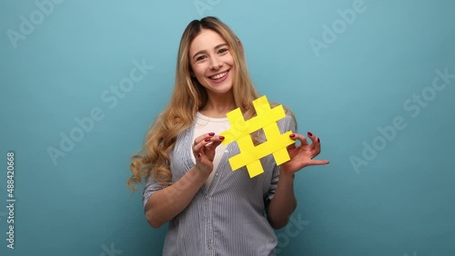 Smiling beautiful woman holding yellow hashtag symbol, making important topic popular, setting trends on Internet, wearing striped shirt. Indoor studio shot isolated on blue background.