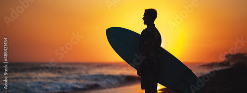 Canvas Print Silhouette of surfer in hawaiian t-shirt with surf board standing on the beach a