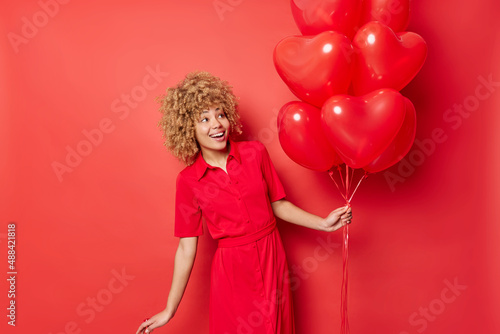 Positive curly haired young woman wears dress poses with big bunch of inflated heart balloons celebrates Valentines Day isolated over vivid red backgrund. Special occasion and celebation concept