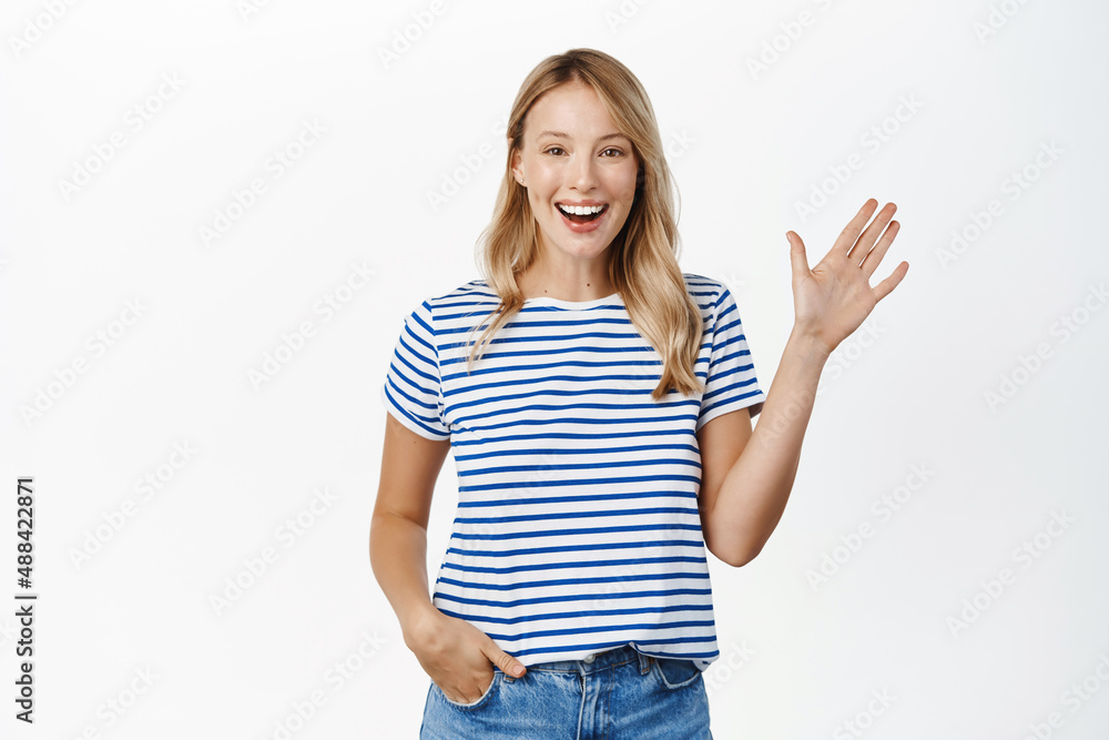 Happy and friendly blond girl smiling, waving hand and saying hello, hi gesture, greeting you with carefree relaxed face expression, white background Happy and friendly blond girl smiling, waving hand and saying hello, hi gesture, greeting you with carefree relaxed face expression, white background