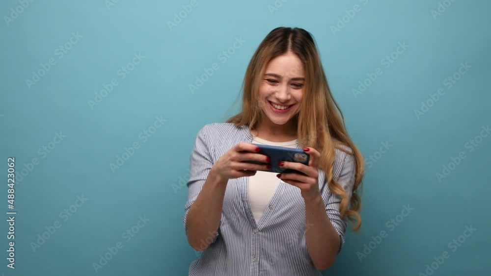 Adorable happy positive woman using mobile phone with attentive focused expression, playing video game on cellphone, wearing striped shirt. Indoor studio shot isolated on blue background. Adorable happy positive woman using mobile phone with attentive focused expression, playing video game on cellphone, wearing striped shirt. Indoor studio shot isolated on blue background.