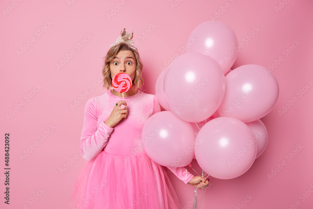 Surprised young woman looks wondered at camera covers mouth with heart shaped caramel candy wears festive dress holds bunch of helium balloons isolated over pink background. Festive occasion