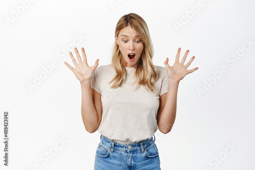 Surprised young beautiful woman shaking hands, screaming and looking down at surprise, gift or promo text below, standing over white background