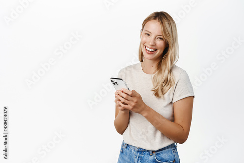 Cellular technology, mobile apps concept. Smiling beautiful woman using smartphone, looking at her phone with happy face, reading, standing over white background