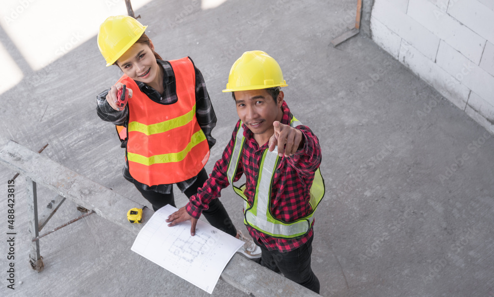 Fototapeta premium Top view of two Asian civil engineers wearing safety hardhat standing looking blueprint and pointing while checking infrastructure in the construction site. 
