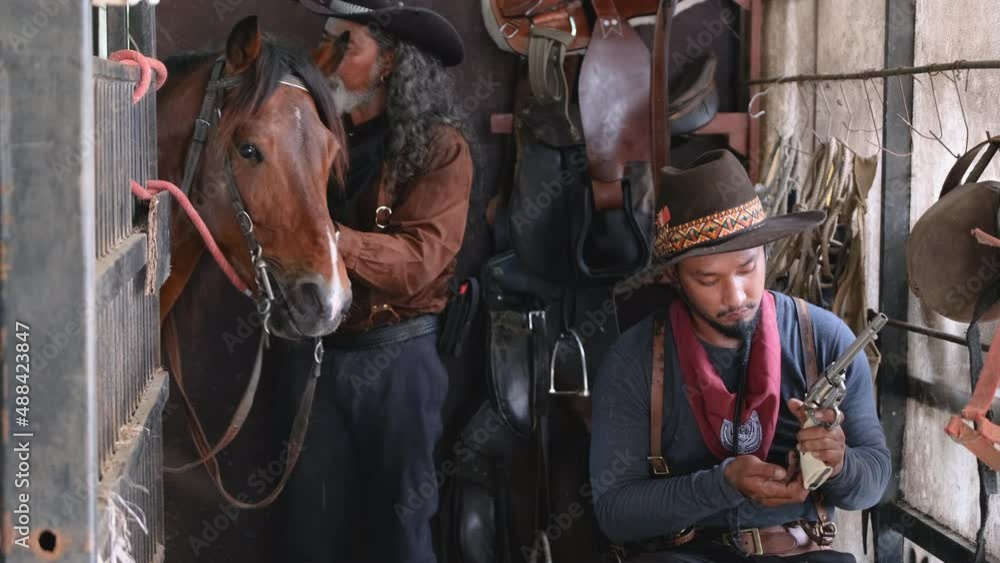 Two man with cowboy costume stay in horse stable that one check the bullet of short gun and other one take care horse in the back. Two man with cowboy costume stay in horse stable that one check the bullet of short gun and other one take care horse in the back.