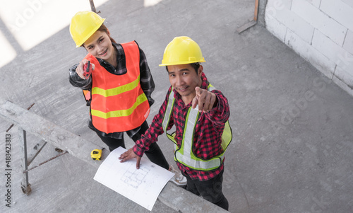 Top view of two Asian civil engineers wearing safety hardhat standing looking blueprint and pointing while checking infrastructure in the construction site. 