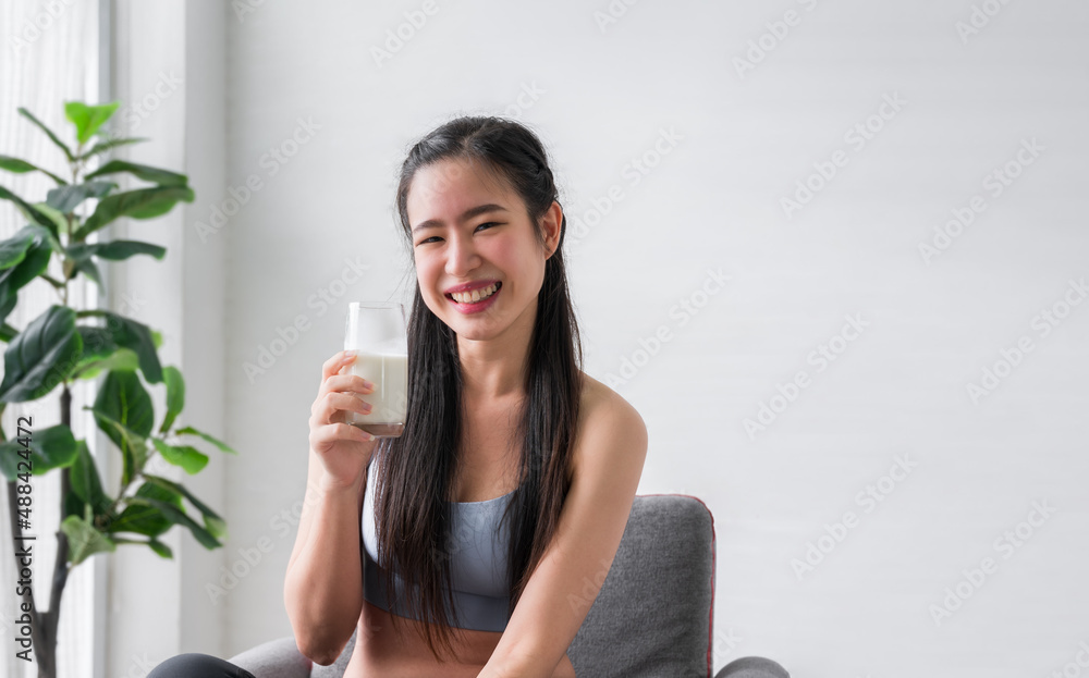 Happy Asian sports girl in sportswear sitting holding and drinking pure milk from glass after exercise in home. Happy Asian sports girl in sportswear sitting holding and drinking pure milk from glass after exercise in home.