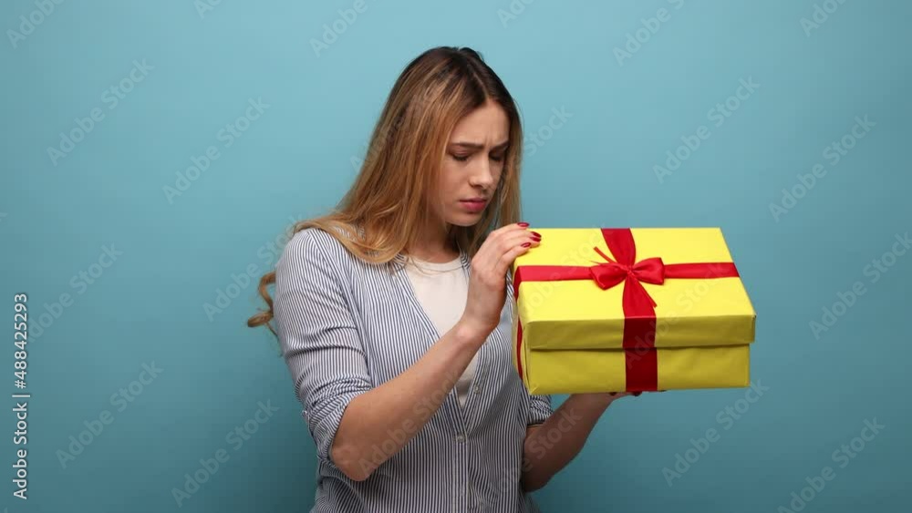 Portrait of upset displeased young adult woman holding gift box, opening present with dissatisfied expression, wearing striped shirt. Indoor studio shot isolated on blue background. Portrait of upset displeased young adult woman holding gift box, opening present with dissatisfied expression, wearing striped shirt. Indoor studio shot isolated on blue background.