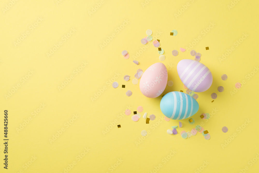 Top view of three pastel painted Easter eggs with cute confetti on the isolated yellow background blank space Top view of three pastel painted Easter eggs with cute confetti on the isolated yellow background blank space