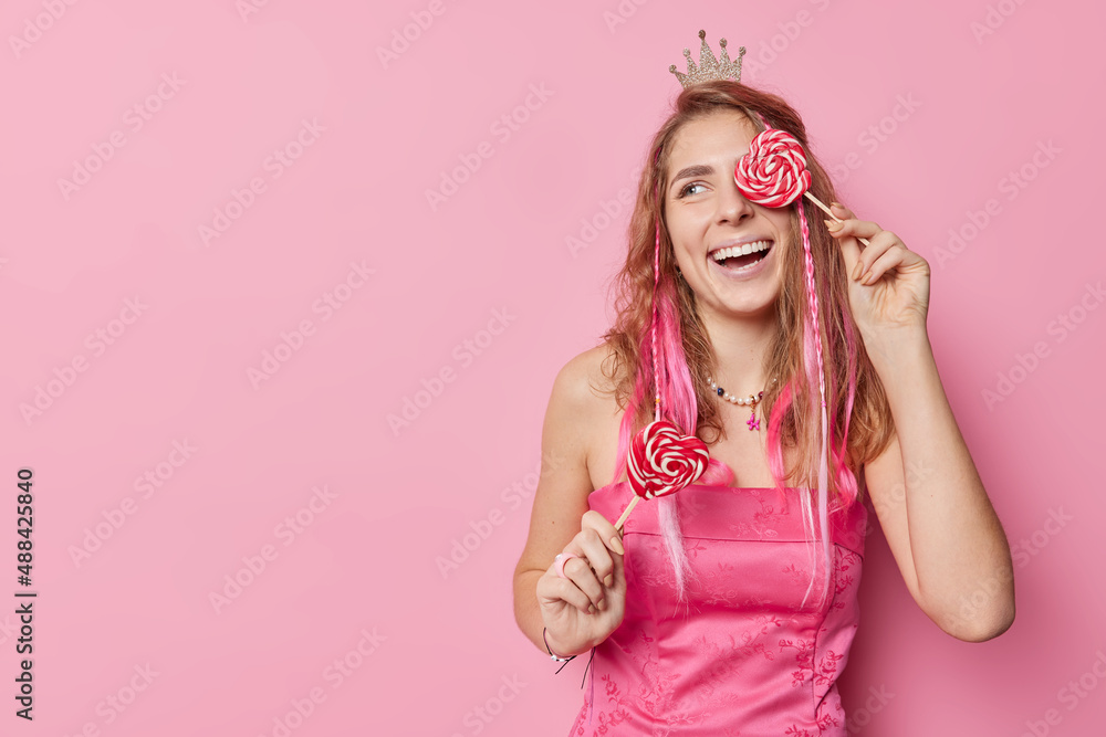 Positive carefree woman covers eye with heart shaped lollipop smiles gladfully wears small crown and dress looks away isolated over pink background with copy space for your promotional content Positive carefree woman covers eye with heart shaped lollipop smiles gladfully wears small crown and dress looks away isolated over pink background with copy space for your promotional content