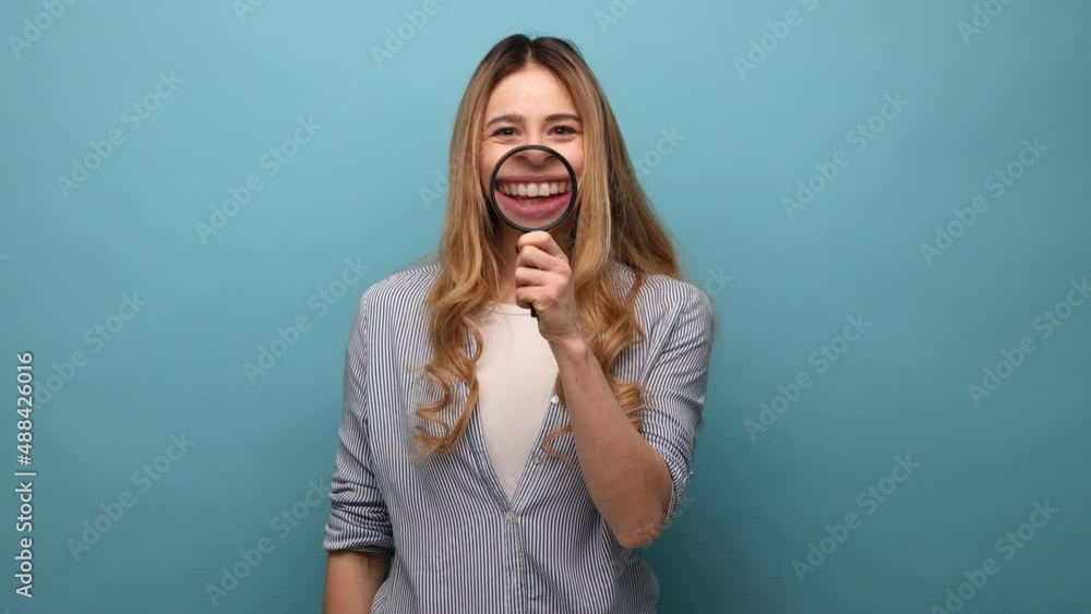 Smile, zoom, tooth care. Portrait of young woman standing and holding magnifying glass on her teeth, looking at camera, wearing striped shirt. Indoor studio shot isolated on blue background. Smile, zoom, tooth care. Portrait of young woman standing and holding magnifying glass on her teeth, looking at camera, wearing striped shirt. Indoor studio shot isolated on blue background.