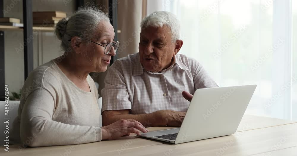 Happy retired couple sit at table looking at laptop discussing new application or social media news, consider online purchase, spend time at home use modern tech, enjoy easy comfort e-services concept Happy retired couple sit at table looking at laptop discussing new application or social media news, consider online purchase, spend time at home use modern tech, enjoy easy comfort e-services concept