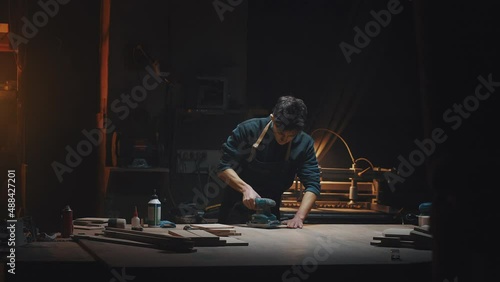 General plan cinematic video of a carpenter at work in a carpentry workshop. The guy polishes wooden parts with a grinder. Dark key, male hobby, family carpentry business photo