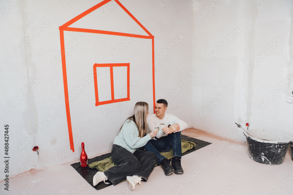 Couple in unfinished building celebrating moving to a new flat. Couple drinking champagne - cheers. Young couple in new property dreaming about renovation near red drawn house on a wall