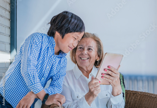 a light-haired caucasian grandmother and her dark-haired asian grandson play with a cell phone on the terrace. they laugh and show happiness. adoption and family concept.
