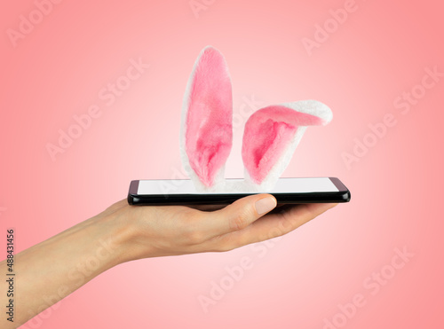 Mobile phone is lying on female palm, Easter Bunny ears sticking out of smartphone screen. Soft pink background. Happy Easter Concept.