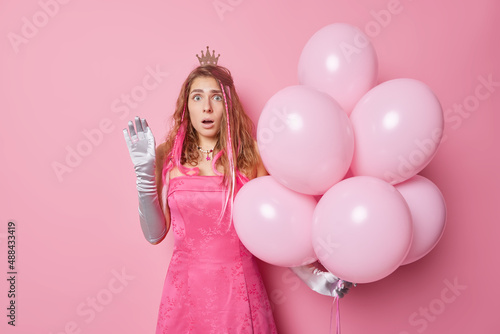 Shocked stunned young woman wears festive dress and gloves holds big bunch of helium balloons comes on birthday party reacts on something amazing isolated over pink background. Holiday concept