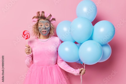 Impressed shocked young woman stares bugged eyes applies hair roller and mud mask holds sweet candy on stick and bunch of inflated balloons prepares for holiday isolated over pink background