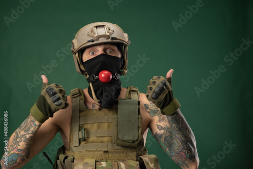 a soldier man in military clothes helmet with a bdsm gag in his mouth expresses emotions, photo joke