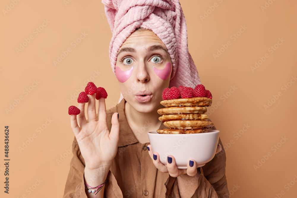Horizontal shot of surprised young European woman stares wondered poses with yummy pancakes puts on raspberries on fingers applies hydrogel patches under eyes dressed in nightwear poses indoor Horizontal shot of surprised young European woman stares wondered poses with yummy pancakes puts on raspberries on fingers applies hydrogel patches under eyes dressed in nightwear poses indoor