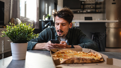 One young caucasian man sitting at the table with pizza order holding mobile phone checking his online bet ticket or crypto currency exchange rate profit winning or loss real people front view