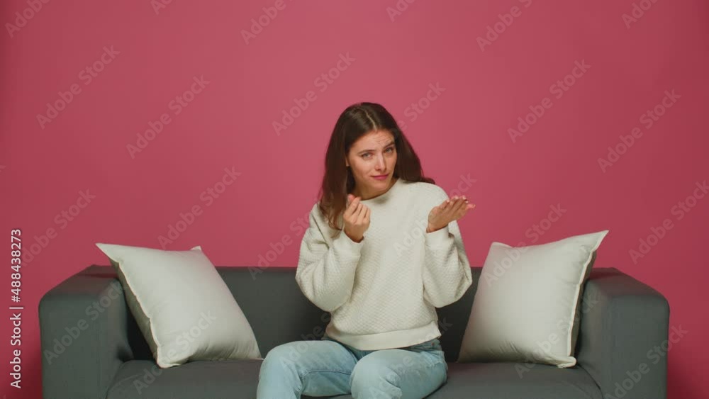 Young girl makes money gesture with hands, asking for salary payment, donation, debt, sitting on modern sofa Young girl makes money gesture with hands, asking for salary payment, donation, debt, sitting on modern sofa