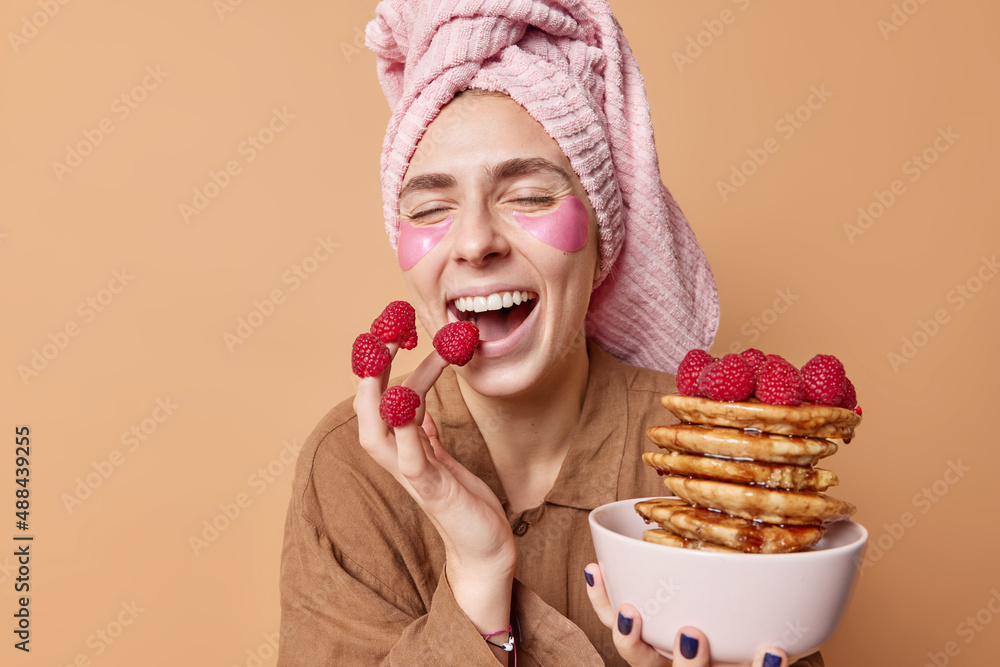 Cheerful happy young woman eats tasty pancakes with raspberry enjoys delicious breakfast has upbeat mood in morning dressed in nightwear wrapped towel on head applies patches to remove puffiness Cheerful happy young woman eats tasty pancakes with raspberry enjoys delicious breakfast has upbeat mood in morning dressed in nightwear wrapped towel on head applies patches to remove puffiness