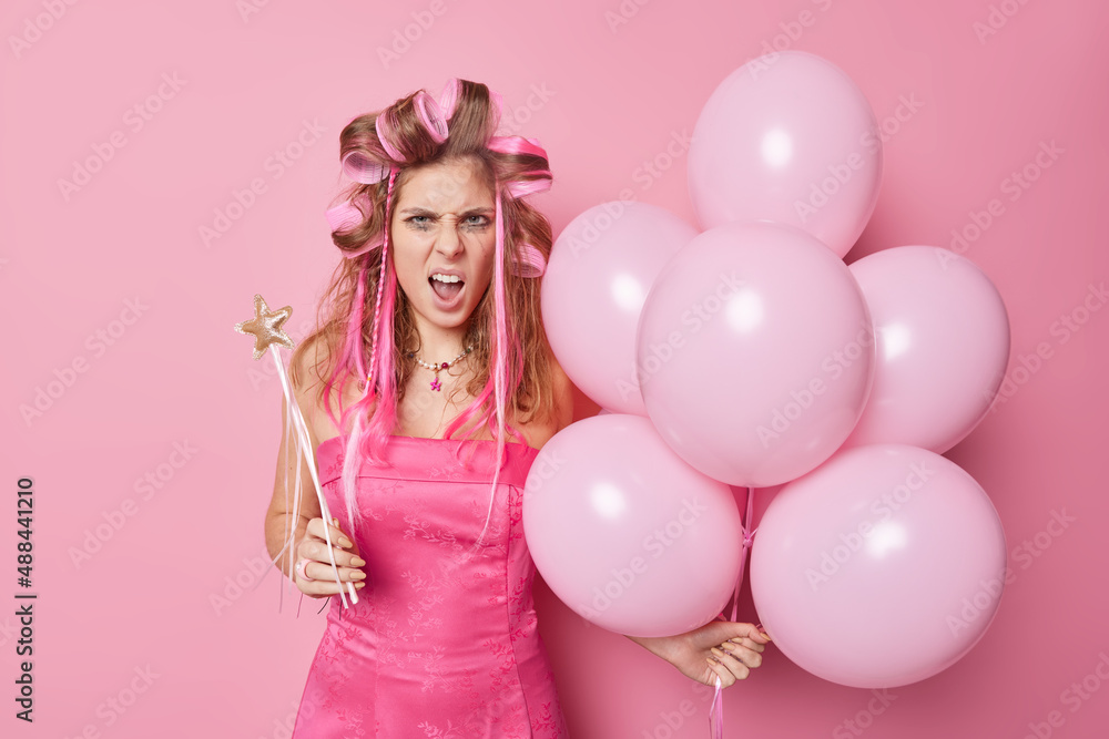 Outraged woman with spoiled makeup exclaims negatively keeps mouth opened holds magic wand and inflated balloons being annoyed by something makes hairstyle prepares for party birthday celebration