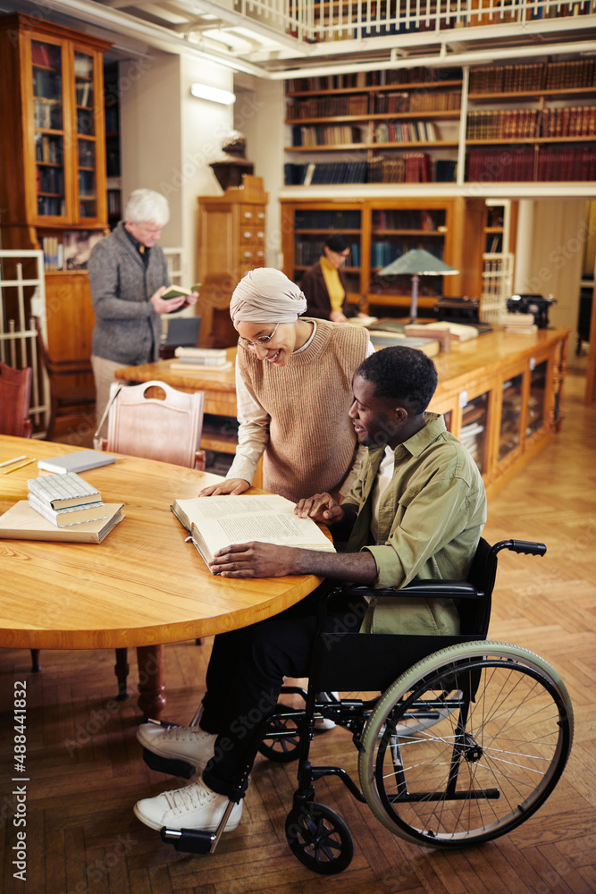 Vertical full length portrait of young African-American using wheelchair in classic college library and reading books with friend Vertical full length portrait of young African-American using wheelchair in classic college library and reading books with friend