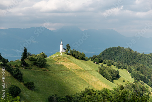 Beautiful landscape of church Jamnik in Slovenia on green hill with blue cloudy sky and mountains background