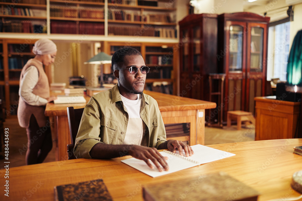 Portrait of young black man with vision impairment reading book in braille at table in college library