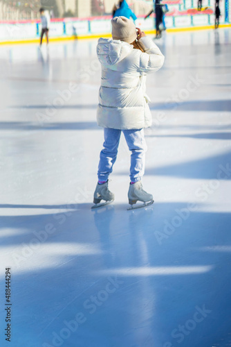Rear view of girl in white tracksuit ice skating on skating rink outside. Healthy winter outdoor activities