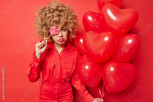 Discontent curly haired woman feels frustrated and lonely on Valentines Dy holds heart balloons covers eye with delicious candy wears dress isolated over vivid red background. Bad mood celebration © wayhome.studio 