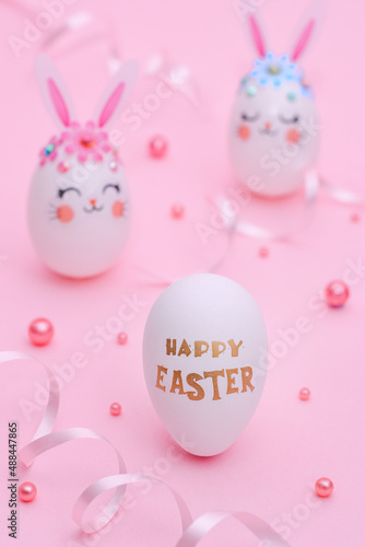 Happy Easter composition on a pink background. White eggs decorated with pink rabbit ears, flowers and muzzles of rabbits. Easter greeting card. Golden greetings text on a white egg: Happy Easter