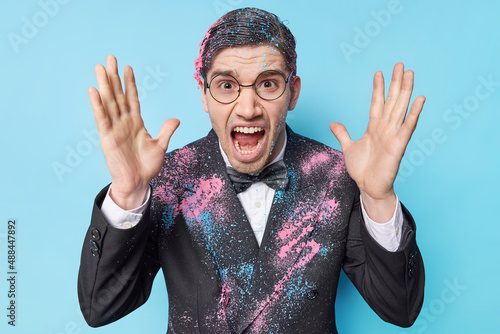 Emotional outraged adult man wears festive suit smeared with coloorful spray shouts loudly keeps palms raised being angry with someone isolated over blue background. Negative emotions concept photo