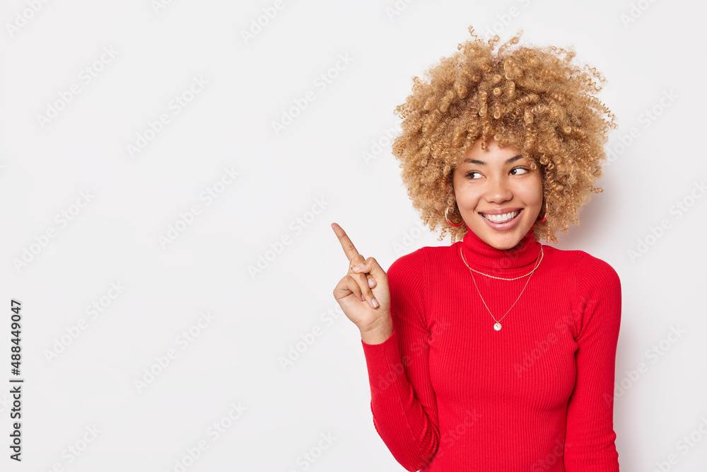 Positive curly haired woman points finger on left side shows blank space for your advertising content wears casual red turtleneck isolated over white background. Place your company title here Positive curly haired woman points finger on left side shows blank space for your advertising content wears casual red turtleneck isolated over white background. Place your company title here
