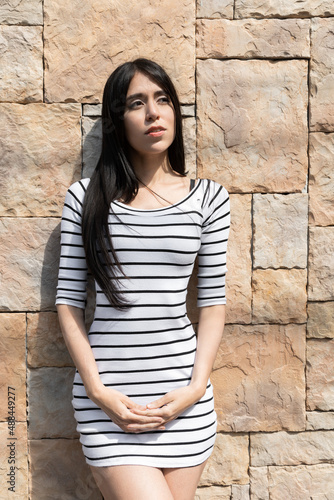 casual style of a young latin woman with long straight hair wearing a striped dress, posing standing on a sunny day with a stone wall in the background, fashion and youthful beauty