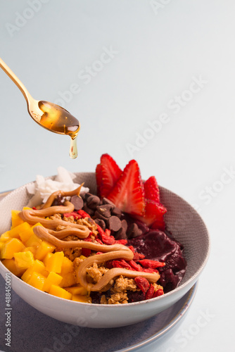serving honey into acai bowl, with strawberries, mango, chocolate chips, coconut, peanut butter. Blue background
