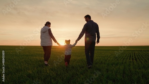 Happy family, dad daughter mom walk together on green grass at sunset. Father mother kid girl walk in park holding hands in sun. Child parents are walking along green field. Family hold hands teamwork