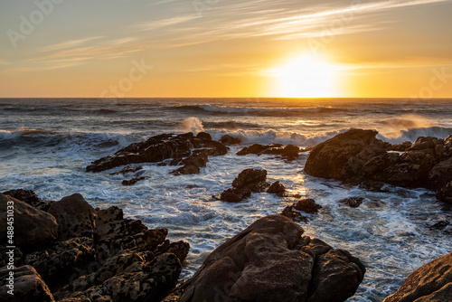 A beautiful orange sunset over the ocean, a bright sun at the edge of the horizon with foaming waves crashing against the dark rocks on the shore