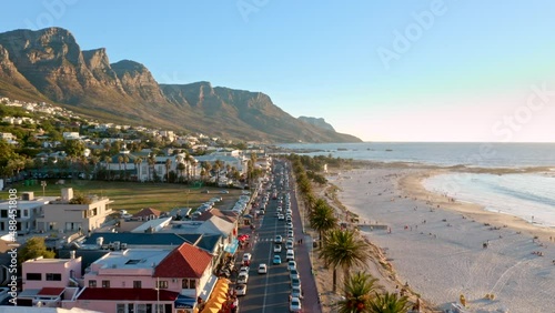 Low altitude aerial shot flying directly overhead the busy main street of Camps Bay's beachfront in Cape Town during sunset with the Twelve Apostles as a backdrop.