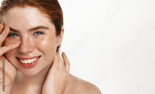 Plus size beauty concept. Curvy redhead model with freckles and white healthy smile. Chubby woman smiling, touching clean, glowing fresh skin without acne, white background