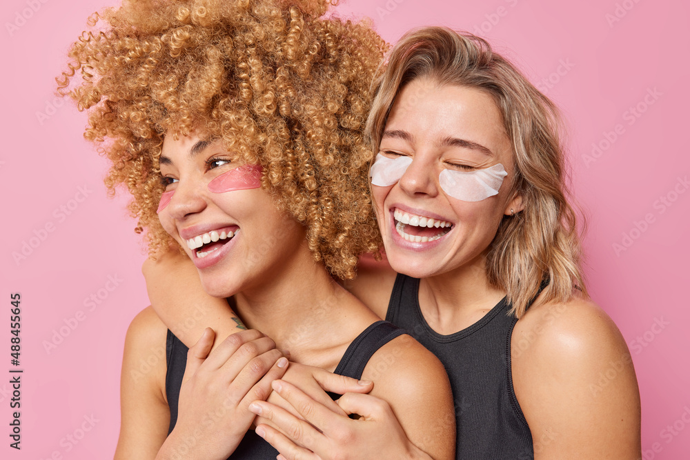 Horizontal shot of cheerful young women embrace and have fun apply beauty hydrogel patches under eyes take care of delicate skin smile broadly wear black t shirts isolated over pink background - obrazy, fototapety, plakaty 