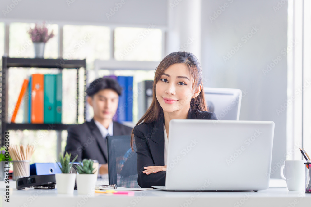 Attractive cheerful young business woman with laptop at office and blurred colleague sitting in the background Attractive cheerful young business woman with laptop at office and blurred colleague sitting in the background