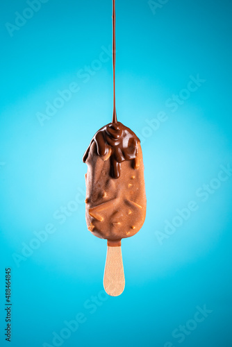 Chocolate popsicle ice cream with almond nuts flavour on wood stick and liquid chocolate falling, isolated on blue background.