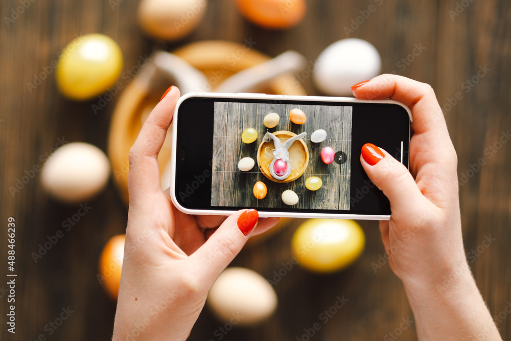 Woman holding mobile phone and making photo of eggs painted for Easter isolated on wooden background. Easter eggs flat lay on rustic table. Easter concepts Woman holding mobile phone and making photo of eggs painted for Easter isolated on wooden background. Easter eggs flat lay on rustic table. Easter concepts